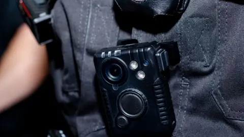 Getty Images Close-up of police body camera attached to an officer's uniform. The device is a small black rectangle-shaped box. The officer's uniform is black.