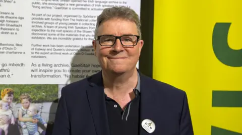A man standing in front of an exhibition display of images and text. He is wearing a blue blazer, polo shirt and glasses. He has short greying hair. 