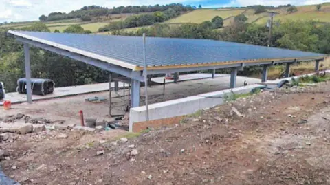 Bradford Council A partially built structure with a large sloped metal roof supported by steel beams, surrounded by construction materials on an unfinished ground, set against rolling hills and fields under a partly cloudy sky.