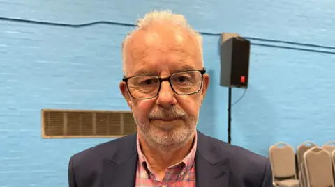A man wearing glasses, a red shirt with a dark-coloured jacket. He is stood in a sports hall with a blue wall. There are chairs and a speaker in the background.