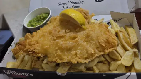 Leanne Jones A tray of battered fish and chips with a slice of lemon and a pot of mushy peas. Hikary's can be seen on the side of the tray.
