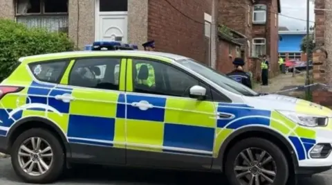 BBC: A police car is parked on the other side of a cobbled alley. Four police officers can be seen blocking the entrance to the alley with police tape.