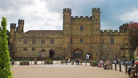 The front of an abbey. There are people sitting outside it.