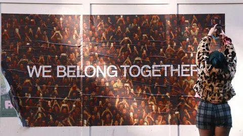 Getty Images Una donna tiene la fotocamera del telefono in alto sopra la testa per scattare una foto di Harry Styles "Apparteniamo insieme" poster su un muro a Soho, Londra.