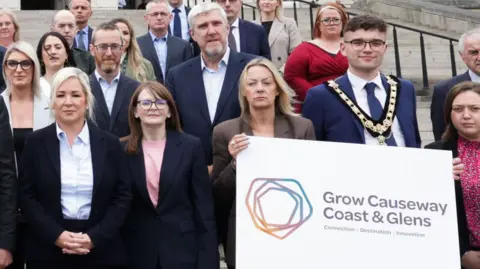 Michelle O'Neill,  Caoimhe Archibald and John O'Dowd stood on the steps of Stormont with members of Causeway Coast & Glens 