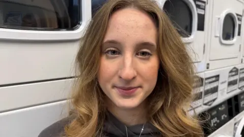 BBC/Alice Lilley Erin is a second-year child nursing student. In this image she is smiling in front of the washing machines in a laundry room on campus at the University of York. Erin is wearing a grey top and has light brown hair. 
