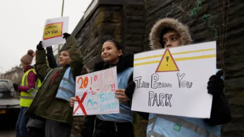 Four year 6 students wearing coats stood by the road with road safety signs 
