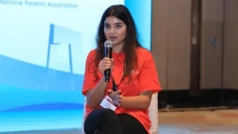 A woman with dark brown long hair and a bright orange tshirt sits on a chair on a stage holding a microphone to her mouth. She's wearing a white pass on a lanyard. Behind is a big screen with a blue background.