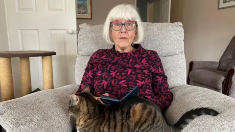 Liz Walton is pictured sitting in an armchair wearing a pink and red patterned blouse with a tabby cat sitting on her lap. She has short white hair with a fringe. Walton is wearing glasses with blue and pink frames.