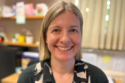 Dr Sarah is wearing a black and cream patterned dress with a black cardigan. She has shoulder length blonde hair and is smiling at the camera. She is in a doctor's surgery with a desk and shelves in the background.