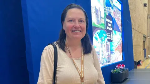 Clare Worden/BBC Alexandra Kemp standing in the hall at LynnSport. She is standing next to a stage and is looking directly at the camera and smiling.
