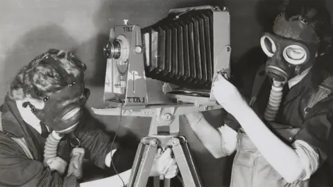Historic England Archive Two Women's Auxiliary Air Force (WAAF) trainees practising photography whilst wearing gas masks to simulate working under active service conditions. The photograph was taken during training at No.2 School of Photography in Blackpool