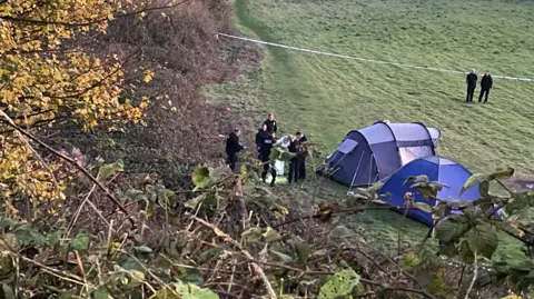BBC Eight uniformed police officers on a field next to two tents, which are blue and on a large grass field. On the left are huge trees and shrubbery.