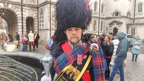 Brian Wilson has a salt and pepper beard and is wearing a beefeater type hat with a tartan scarf across his chest. He is holding mace and is wearing a black and red uniform