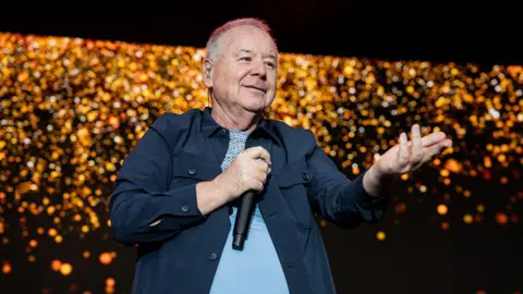 Getty Images A man with short grey hair is holding a microphone on stage