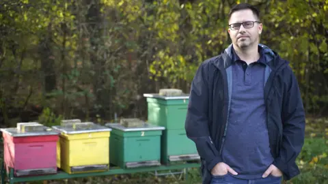 Martin Bystriansky Wearing a blue jacket and blue shirt, Dr Juraj Majtán stands in front of four beehives.
