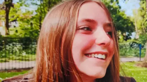 Close up of the face and head of a young girl. She has red hair and is smiling. In the background is a park.