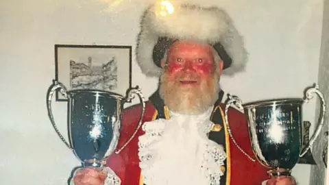 Trevor Heeks Trevor Heeks, who has a red face and big grey beard is wearing his ceremonial outfit with a furry hat, red coat and white ruff. He is holding two large silver trophies