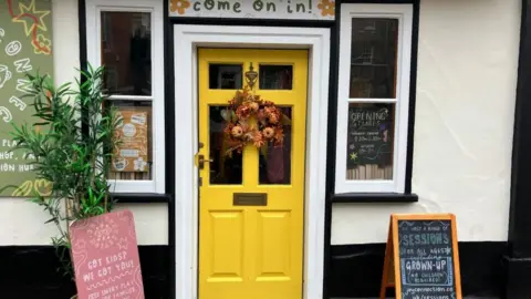 Vikki Irwin/BBC The photo shows a bright yellow door decorated with an autumn wreath. Above the door is a sign to say Come on in. Two chalkboard signs advertising sessions and activities are either side of the door. 