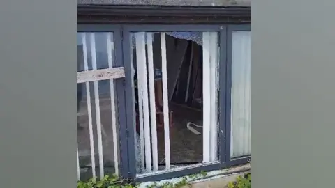 Lance Martin A close-up of black framed three-pane window with the middle pane smashed, showing through into a room beyond