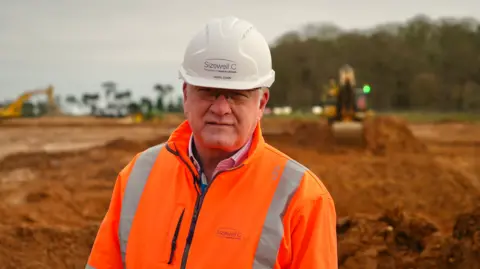 Martin Giles/BBC Nigel Cann stands within construction site and smiles at the camera. He wears a white hard helmet and orange hi-vis jacket. Construction vehicles can be seen behind him. 