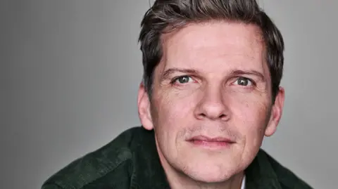 Ruth Crafer Nigel Harman looks directly at the camera. He is wearing a dark green jacket and has short, dark brown hair and green eyes.
