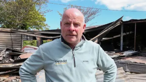 A man wearing a blue jumper stands in the wreckage of a fire. he has his hands on his hips and stares into the camera. There is metal sheeting lying on the ground.