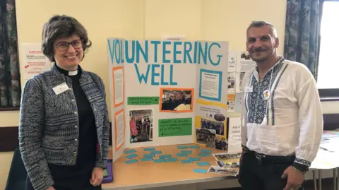 Jon Wright/BBC Reverend Sally Gaze and Romeo Mustata stand either side of a display board with signs about volunteering