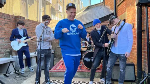 A group of musicians from the band EMF playing on a sunny day in Cinderford and smiling.