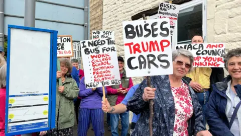 A crowd of people holding up placards in front of a bus stop, campaigning against the scrapping of the 84/85 bus service