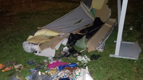 Various items of waste seen scattered on a grass area. There appears to be a sofa as well as a mirror and several food packets.