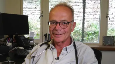 A photo of Dr Simon Opher sat at his desk wearing glasses and a stethoscope around his shoulders