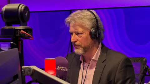 Mike Sheridan is wearing a black suit with a pink shirt underneath. He has a pair of wired black headphones on and is sat inside a radio studio with purple screens. He is staring in front of him towards the presenter.
