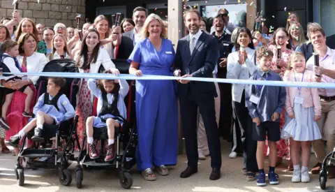 Sir Gareth Southgate, wearing a dark suit, white shirt and patterned tie, cuts a ribbon to open The Build project at Martin House in Boston Spa, surrounded by staff, families and children.
