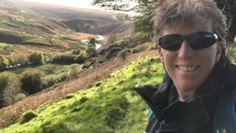 Sue Applegate Woman with short blond hair and dark glasses standing on a hillside in Exmoor National Park, with a valley behind her