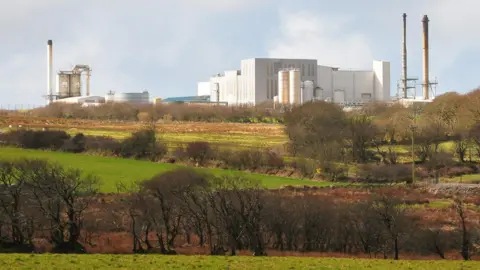 Davidstow Creamery photographed from across a few fields on a sunny day. It is a large white building. There are multiple silos and three large chimneys. In the foreground are fields lined with trees and bushes.
