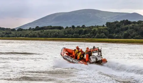 Kim Ayres A lifeboat skipping across the water surface on the Solway estuary with trees and hills in the background