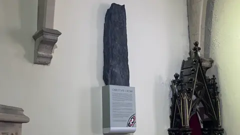 Oruths cross is mounted to a church wall. It's resting on a podium which is titles "Orsuths Cross"
