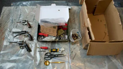 National Crime Agency Three antique handguns, a box of bullets, some screwdrivers and scissors, and a cardboard box laid out on a table 