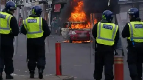 Four police officers with helmets and shields standing in a line in front of a red car on fire.