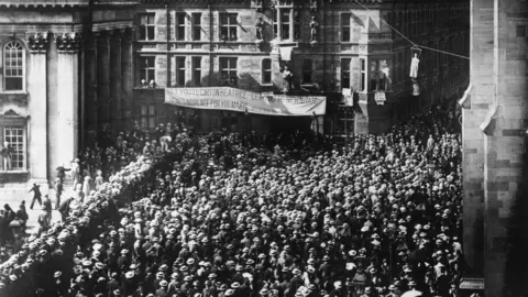 Getty Images A black and white image showing a street packed with men wearing hats in 1897 in Cambridge after the vote to deny women degrees. To the right can just be seen a corner of the Senate House. In the middle is a banner strung off another buildings and on the far right an effigy of a woman is strung up above the crowds.