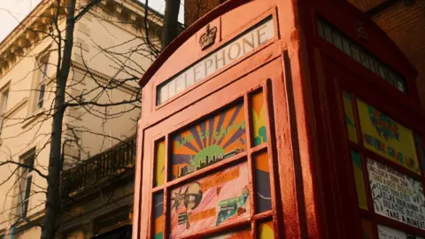 A red phone box with colourful artwork in the glass panes. The box has been transformed into a resource for rough sleepers to use to find support.