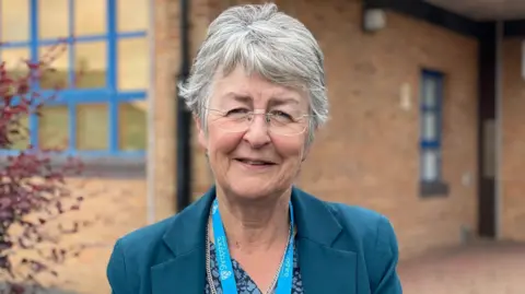 A woman with short grey hair and glasses in a teal jacket outside a brown brick building