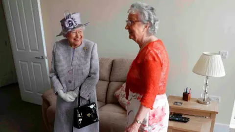PETER NICHOLLS/AFP via Getty Images Queen Elizabeth II standing in a living room with an elderly woman. The Queen is dressed in a formal coat with matching hat and gloves, holding a black handbag. The woman is wearing a bright red cardigan over a floral skirt. They are standing near a beige sofa, with a wooden side table next to it that holds a lamp, a remote control, and some stationery items. 