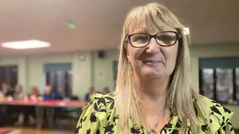 A close-up of Lisa, smiling to camera, wearing a yellow blouse with a black flowery pattern, and she has long blond hair and black-rimmed glasses. Behind her is the cafe, which is blurred.