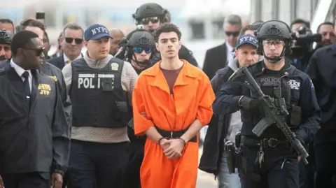 Getty Images Luigi Mangione is in an orange prison jumpsuit which he is walked while handciffed extradited alongside several armed officers. They are outside having just gotten off a helicopter in New York. His face has no expression.