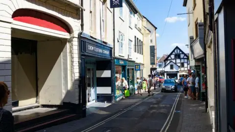 Getty Images A street in Ludlow town centre. There are shops on either side of the narrow road, which has double yellow lines, and people walking on the pavements. The road leads to a Tudor-style black and white building.