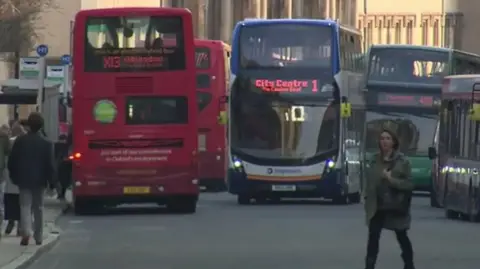 Four buses on a street in central Oxford with a pedestrian walking across the road while other people wait at bus stops