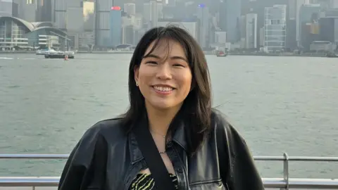 Ines Tan Wearing a black leather jacket. Ines Tan smiles while standing at a waterfront.