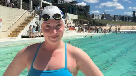 Katie Bain A white woman, Katie, looking at the camera. She is wearing blue swimming suit, blue and white swimming hat and black swimming goggles. The outdoors pool is behind her. 